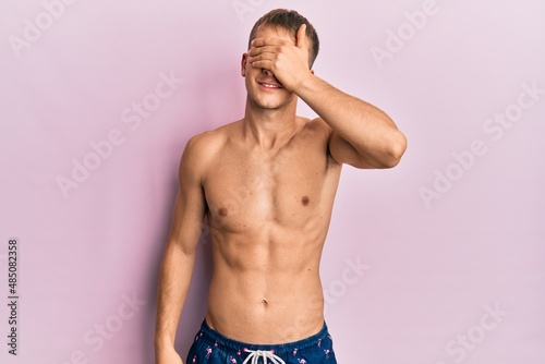 Young caucasian man wearing swimwear smiling and laughing with hand on face covering eyes for surprise. blind concept. photo
