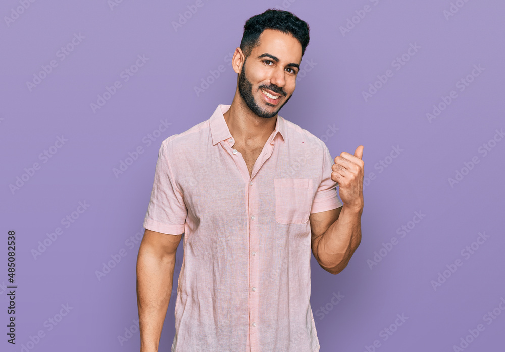 Hispanic man with beard wearing casual shirt smiling with happy face looking and pointing to the side with thumb up. Hispanic man with beard wearing casual shirt smiling with happy face looking and pointing to the side with thumb up.