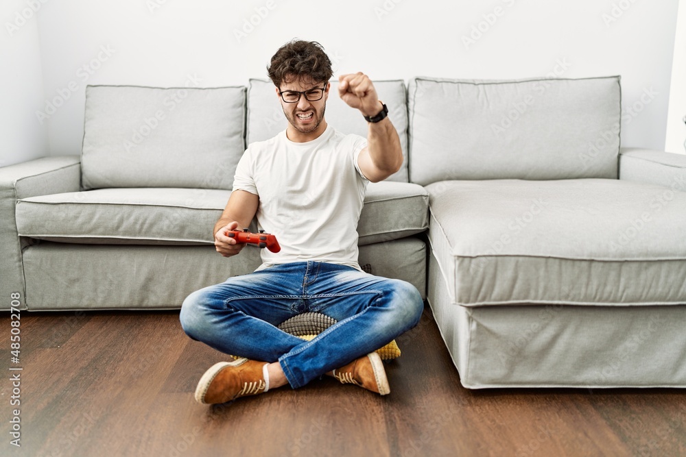 Hispanic man playing video game holding controller by the sofa annoyed and frustrated shouting with anger, yelling crazy with anger and hand raised