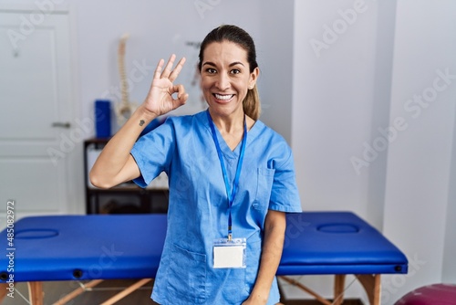 Young hispanic woman wearing physiotherapist uniform standing at clinic smiling positive doing ok sign with hand and fingers. successful expression.