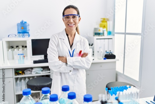 Young hispanic woman wearing scientist uniform with arms crossed gesture at laboratory