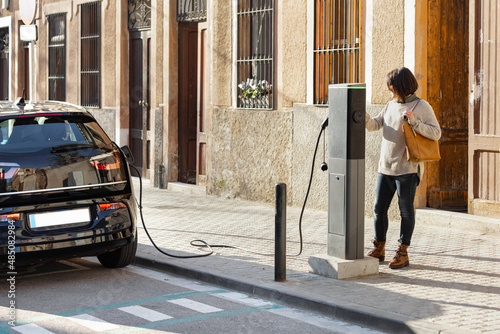 Photography Woman standing near a recharge city station to charge her electric car