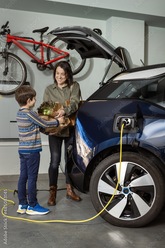 Woman and boy pick up the groceries while electric car is charging at home.