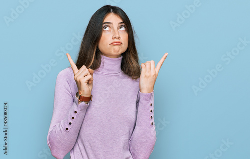 Young beautiful teen girl wearing turtleneck sweater pointing up looking sad and upset, indicating direction with fingers, unhappy and depressed.