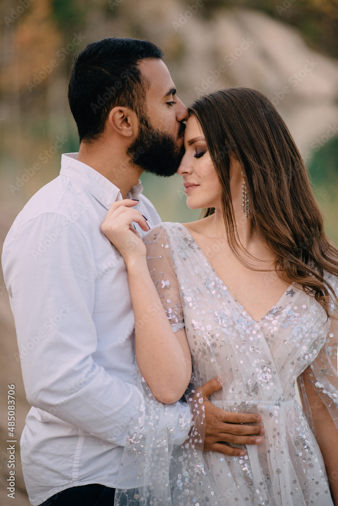 © MZaitsev - Interracial relations. Middle Eastern groom and Caucasian bride hug against a beautiful lake. A man kisses a woman on the cheek. © MZaitsev - Interracial relations. Middle Eastern groom and Caucasian bride hug against a beautiful lake. A man kisses a woman on the cheek.
