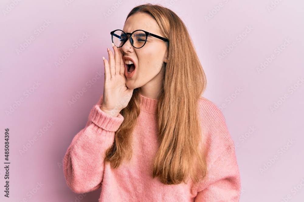 Young blonde woman wearing casual clothes and glasses shouting and screaming loud to side with hand on mouth. communication concept.