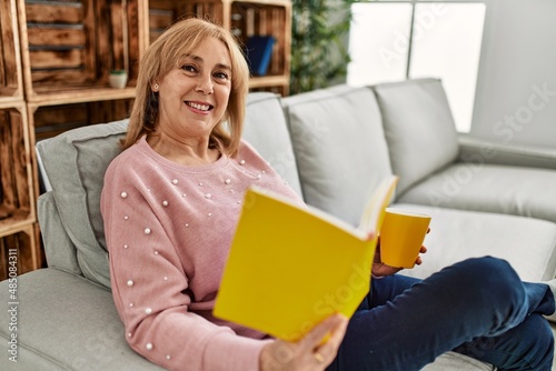 Middle age blonde woman smiling happy reading book and drinking coffee at home.