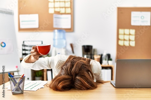 Young hispanic businesswoman overworked with head on table and holding cup of coffee at the office.