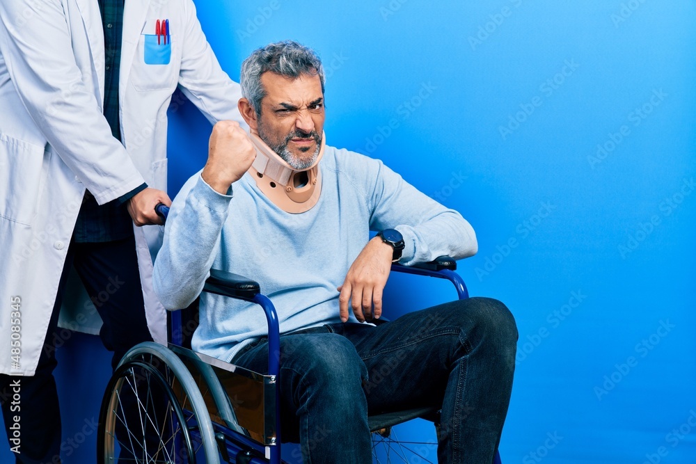 Handsome middle age man with grey hair on wheelchair wearing cervical collar angry and mad raising fist frustrated and furious while shouting with anger. rage and aggressive concept.