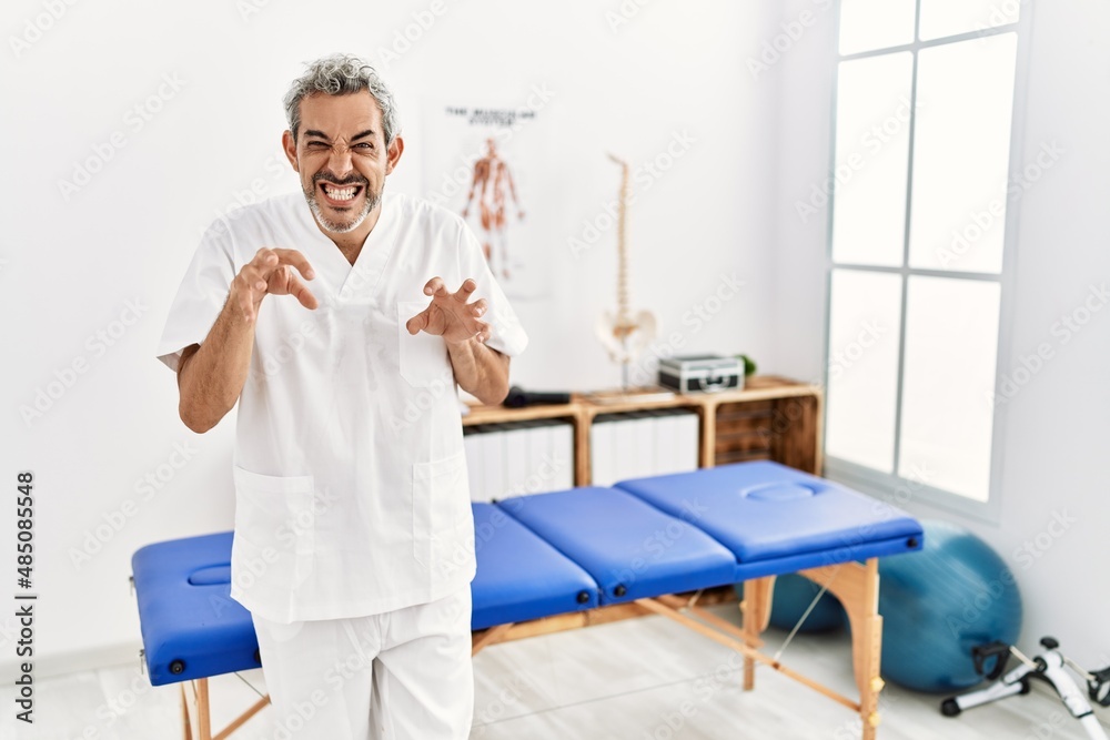 Middle age hispanic therapist man working at pain recovery clinic smiling funny doing claw gesture as cat, aggressive and sexy expression Middle age hispanic therapist man working at pain recovery clinic smiling funny doing claw gesture as cat, aggressive and sexy expression