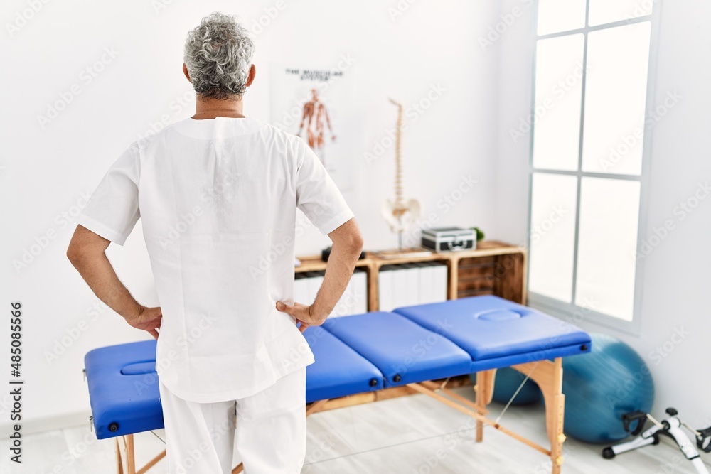 Middle age hispanic therapist man working at pain recovery clinic standing backwards looking away with arms on body