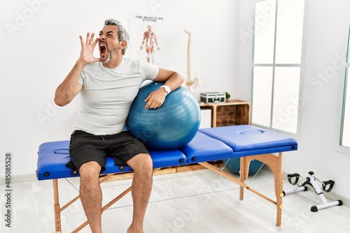 Middle age hispanic man at pain recovery clinic holding pilates ball shouting and screaming loud to side with hand on mouth. communication concept.