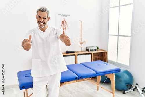 Middle age hispanic therapist man working at pain recovery clinic approving doing positive gesture with hand  thumbs up smiling and happy for success. winner gesture.