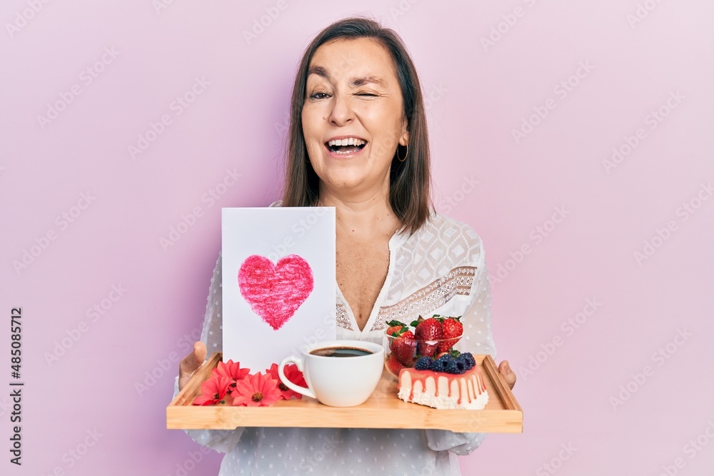 Middle age hispanic woman holding tray with breakfast food and heart draw winking looking at the camera with sexy expression, cheerful and happy face. Middle age hispanic woman holding tray with breakfast food and heart draw winking looking at the camera with sexy expression, cheerful and happy face.