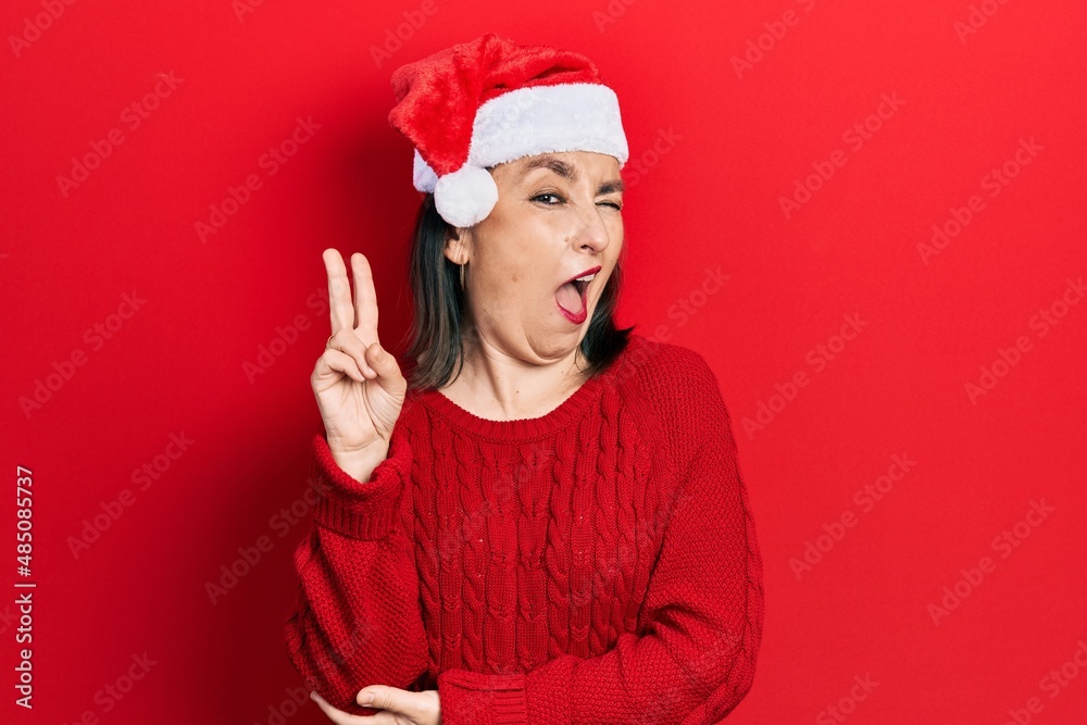 Middle age hispanic woman wearing christmas hat smiling with happy face winking at the camera doing victory sign. number two. Middle age hispanic woman wearing christmas hat smiling with happy face winking at the camera doing victory sign. number two.