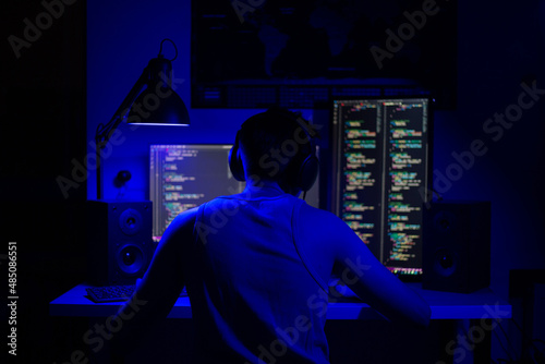 A man sits at a computer in a room at a table at night with blue lighting and programs