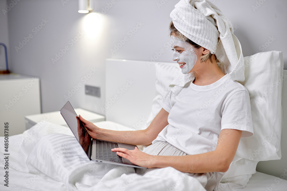 Young attractive woman after shower, with mud facial mask, typing on laptop in bed, working or studying online at cozy bedroom at home. Working online, freelance concept. beauty, skin care concept Young attractive woman after shower, with mud facial mask, typing on laptop in bed, working or studying online at cozy bedroom at home. Working online, freelance concept. beauty, skin care concept