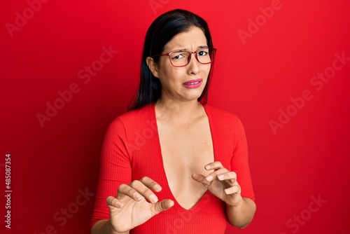 Young latin woman wearing casual clothes and glasses disgusted expression, displeased and fearful doing disgust face because aversion reaction. with hands raised