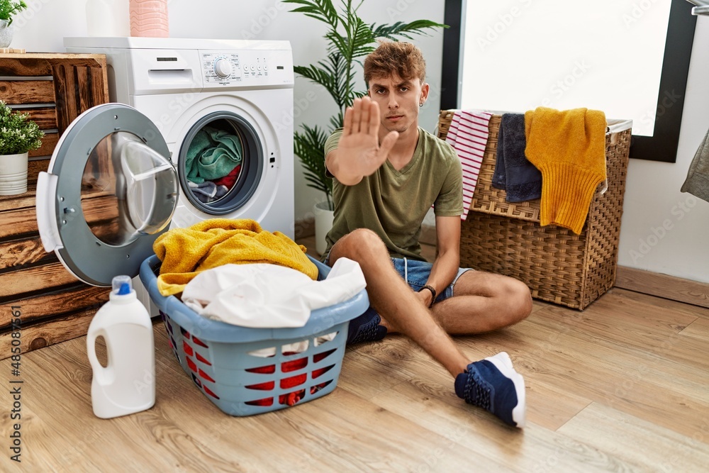 Young caucasian man putting dirty laundry into washing machine doing stop sing with palm of the hand. warning expression with negative and serious gesture on the face. Young caucasian man putting dirty laundry into washing machine doing stop sing with palm of the hand. warning expression with negative and serious gesture on the face.