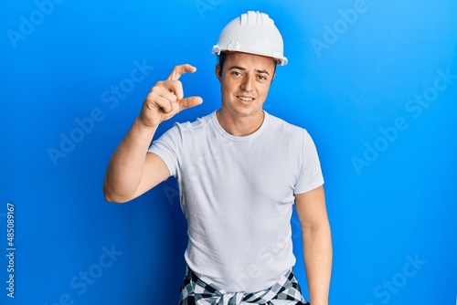 Handsome young man wearing builder uniform and hardhat smiling and confident gesturing with hand doing small size sign with fingers looking and the camera. measure concept.