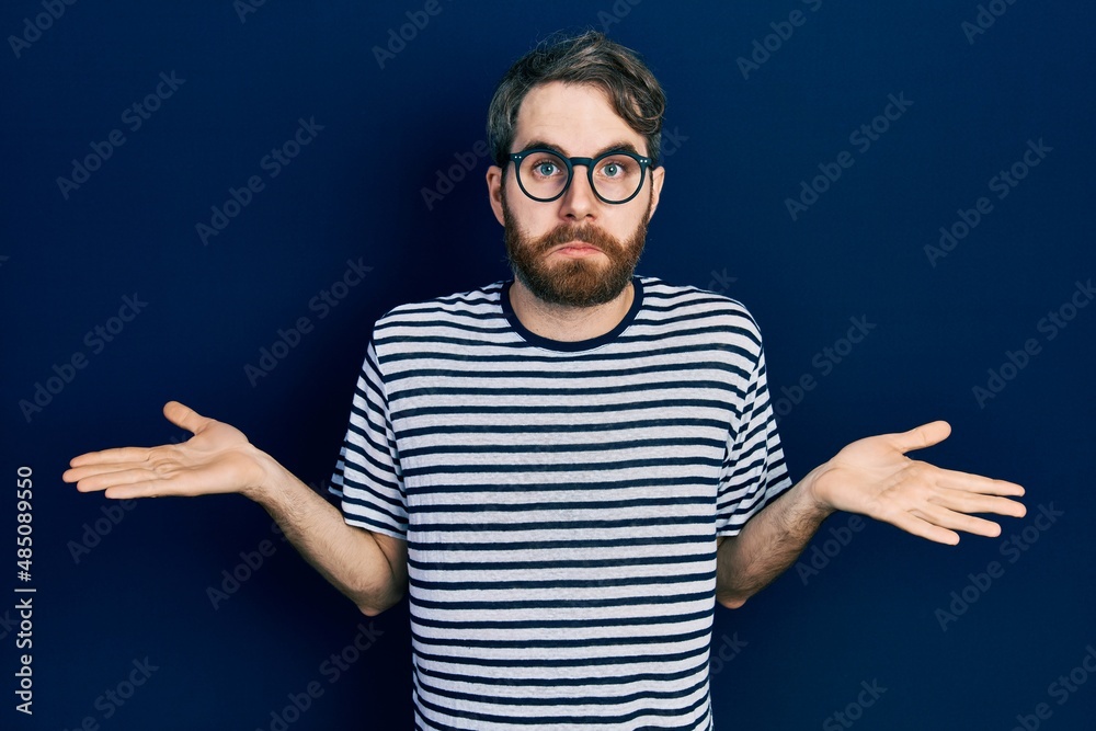 Caucasian man with beard wearing striped t shirt and glasses clueless and confused expression with arms and hands raised. doubt concept. Caucasian man with beard wearing striped t shirt and glasses clueless and confused expression with arms and hands raised. doubt concept.