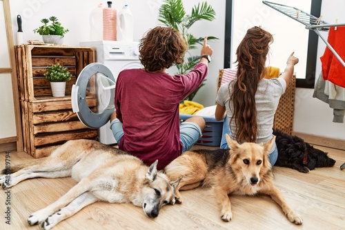 Canvas Print Young hispanic couple doing laundry with dogs posing backwards pointing ahead wi