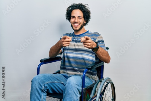 Handsome hispanic man sitting on wheelchair pointing fingers to camera with happy and funny face. good energy and vibes.
