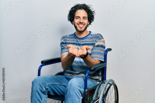 Handsome hispanic man sitting on wheelchair smiling with hands palms together receiving or giving gesture. hold and protection