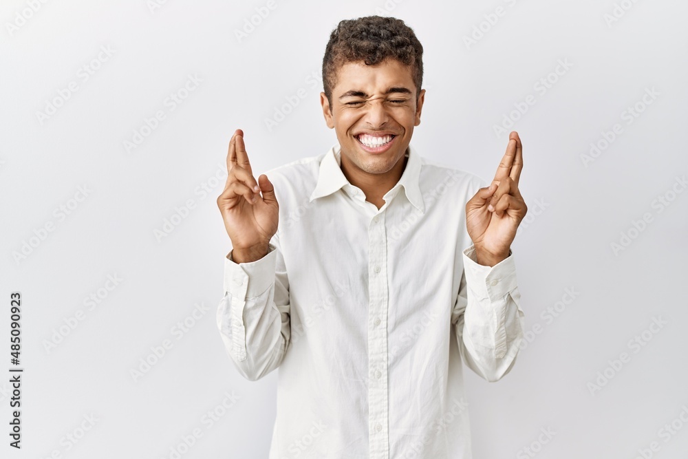 Young handsome hispanic man standing over isolated background gesturing finger crossed smiling with hope and eyes closed. luck and superstitious concept. Young handsome hispanic man standing over isolated background gesturing finger crossed smiling with hope and eyes closed. luck and superstitious concept.
