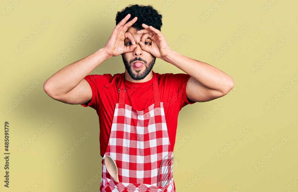 Young arab man with beard wearing cook apron doing ok gesture like binoculars sticking tongue out, eyes looking through fingers. crazy expression. Young arab man with beard wearing cook apron doing ok gesture like binoculars sticking tongue out, eyes looking through fingers. crazy expression.