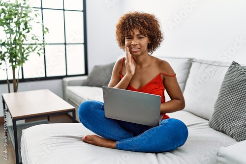 Young african american woman sitting on the sofa at home using laptop touching mouth with hand with painful expression because of toothache or dental illness on teeth. dentist