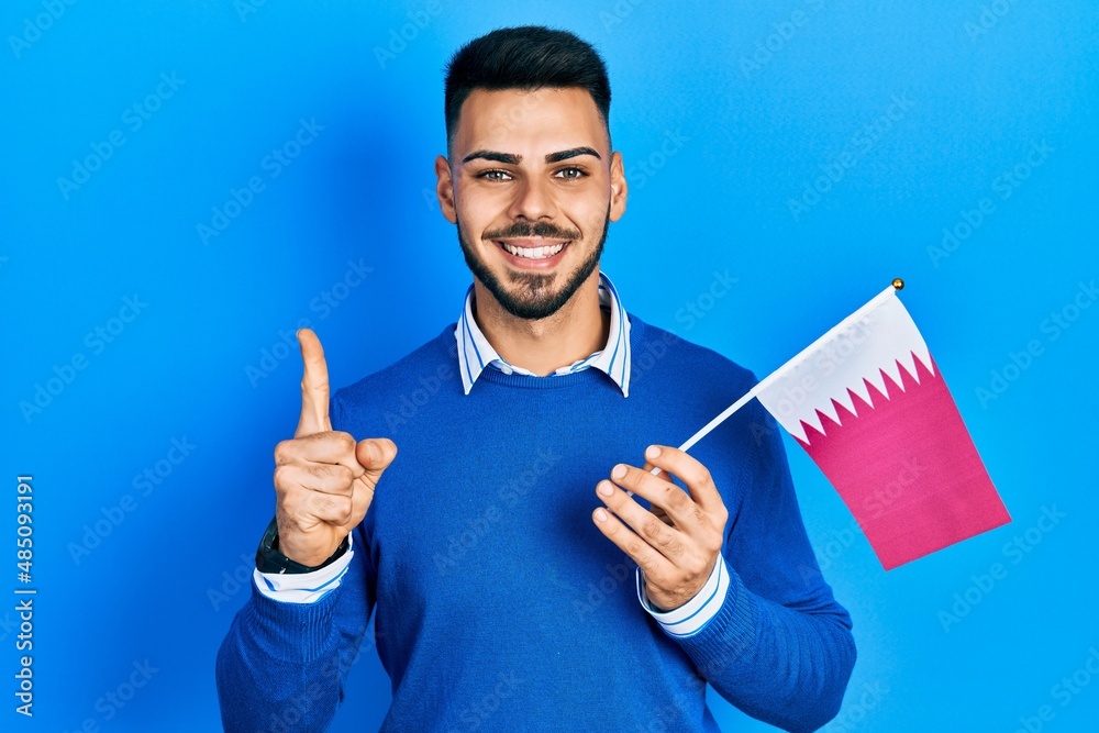 Young hispanic man with beard holding qatar flag smiling with an idea or question pointing finger with happy face, number one Young hispanic man with beard holding qatar flag smiling with an idea or question pointing finger with happy face, number one