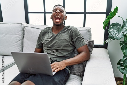 Foto Young african american man using laptop at home sitting on the sofa angry and mad screaming frustrated and furious, shouting with anger