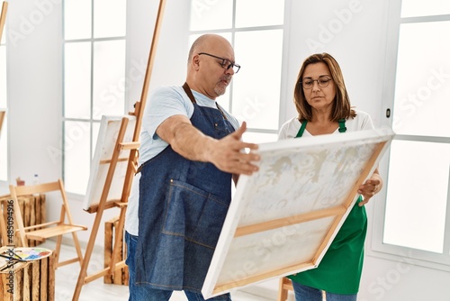 Middle age hispanic painter couple with serious expression looking canvas at art studio.