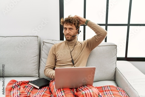 Young handsome man with beard wearing operator headset working from home confuse and wondering about question. uncertain with doubt, thinking with hand on head. pensive concept.