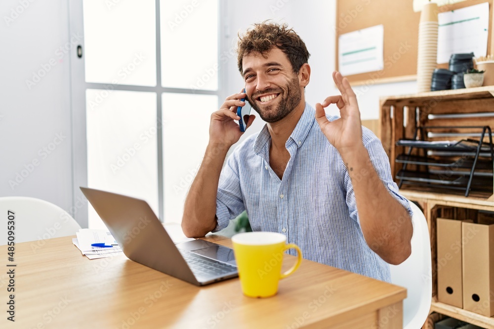 Young handsome man working at the office speaking on the phone doing ok sign with fingers, smiling friendly gesturing excellent symbol Young handsome man working at the office speaking on the phone doing ok sign with fingers, smiling friendly gesturing excellent symbol