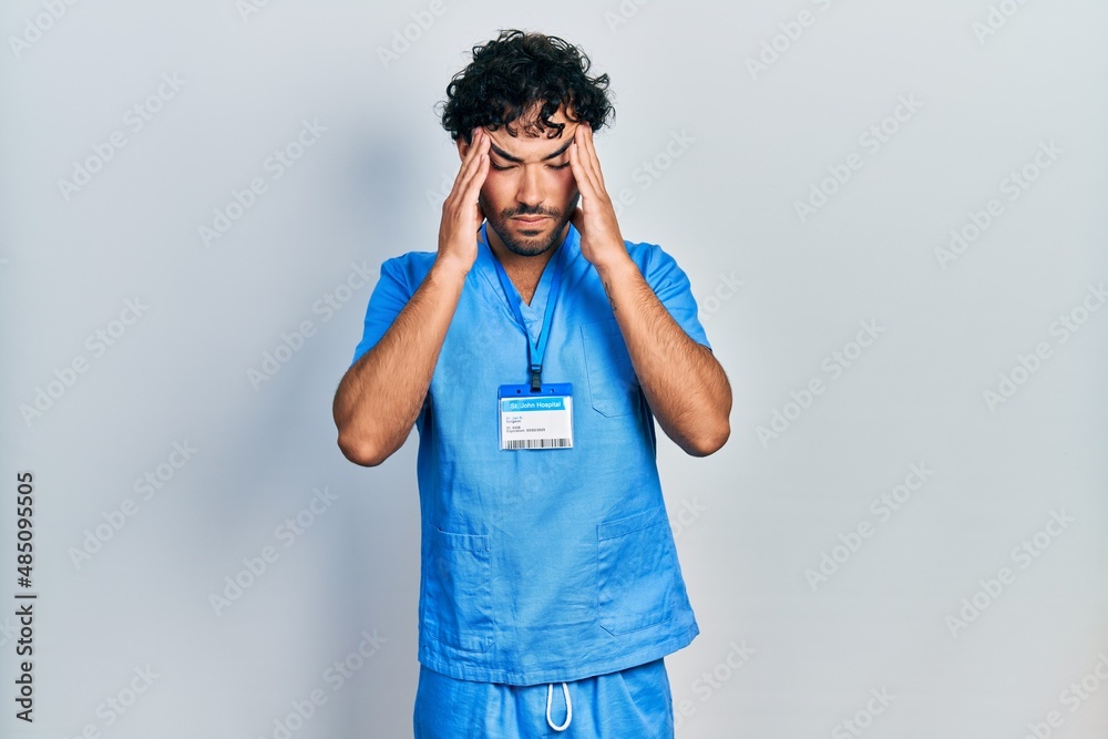 Young hispanic man wearing blue male nurse uniform with hand on head for pain in head because stress. suffering migraine. Young hispanic man wearing blue male nurse uniform with hand on head for pain in head because stress. suffering migraine.