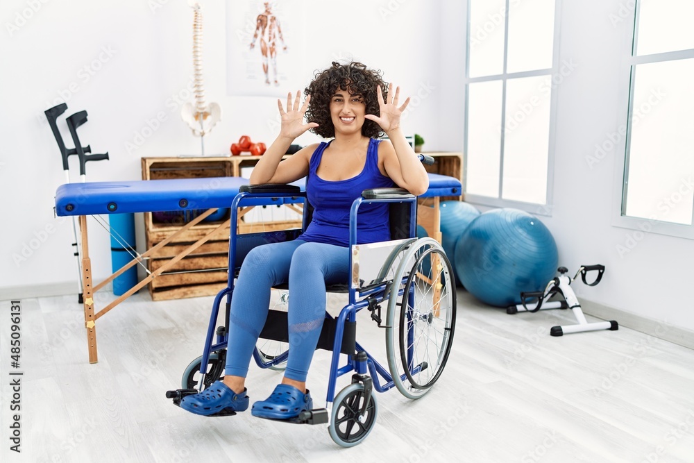 Obraz premium Young middle eastern woman sitting on wheelchair at physiotherapy clinic showing and pointing up with fingers number ten while smiling confident and happy.
