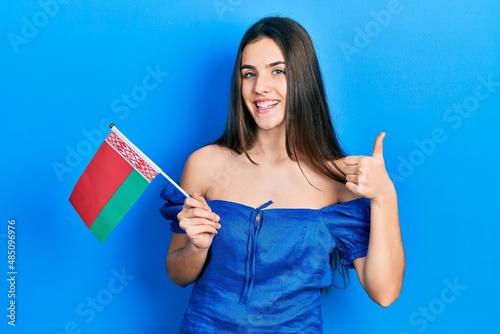Young brunette teenager holding belarus flag smiling happy and positive  thumb up doing excellent and approval sign