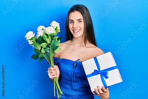 Young brunette teenager holding anniversary present and bouquet of flowers smiling with a happy and cool smile on face. showing teeth.