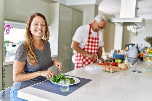 Middle age hispanic woman smiling happy eating salad while man cook at the kitchen.
