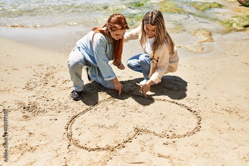 Young lesbian couple of two women in love at the beach. Beautiful women together at the beach in a romantic relationship drawing a heart on the sand