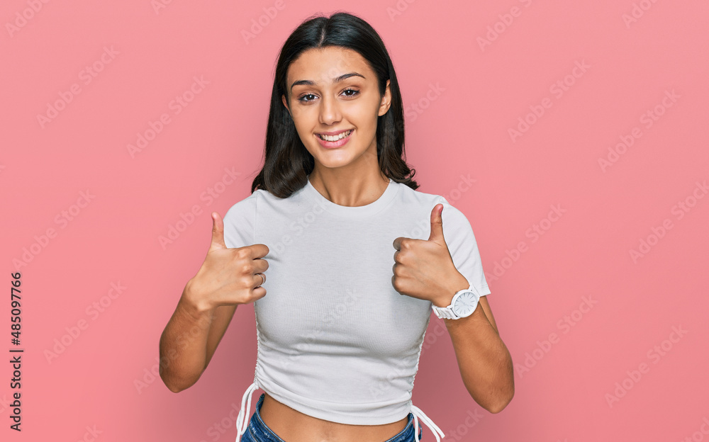 Young hispanic girl wearing casual white t shirt success sign doing positive gesture with hand, thumbs up smiling and happy. cheerful expression and winner gesture.