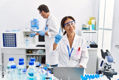 Middle age woman working at scientist laboratory surprised with an idea or question pointing finger with happy face, number one