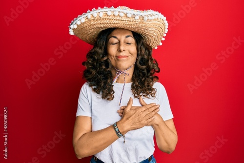 Middle age hispanic woman holding mexican hat smiling with hands on chest with closed eyes and grateful gesture on face. health concept. © Krakenimages.com