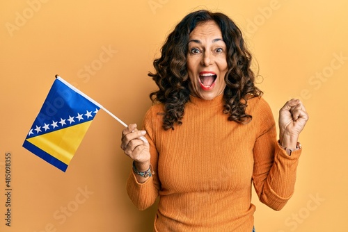 Middle age hispanic woman holding bosnia herzegovina flag screaming proud, celebrating victory and success very excited with raised arm photo