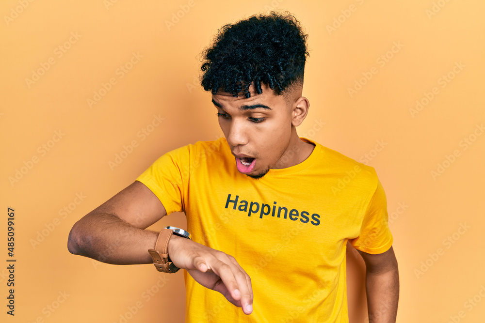 Young african american man wearing t shirt with happiness word message looking at the watch time worried, afraid of getting late Young african american man wearing t shirt with happiness word message looking at the watch time worried, afraid of getting late