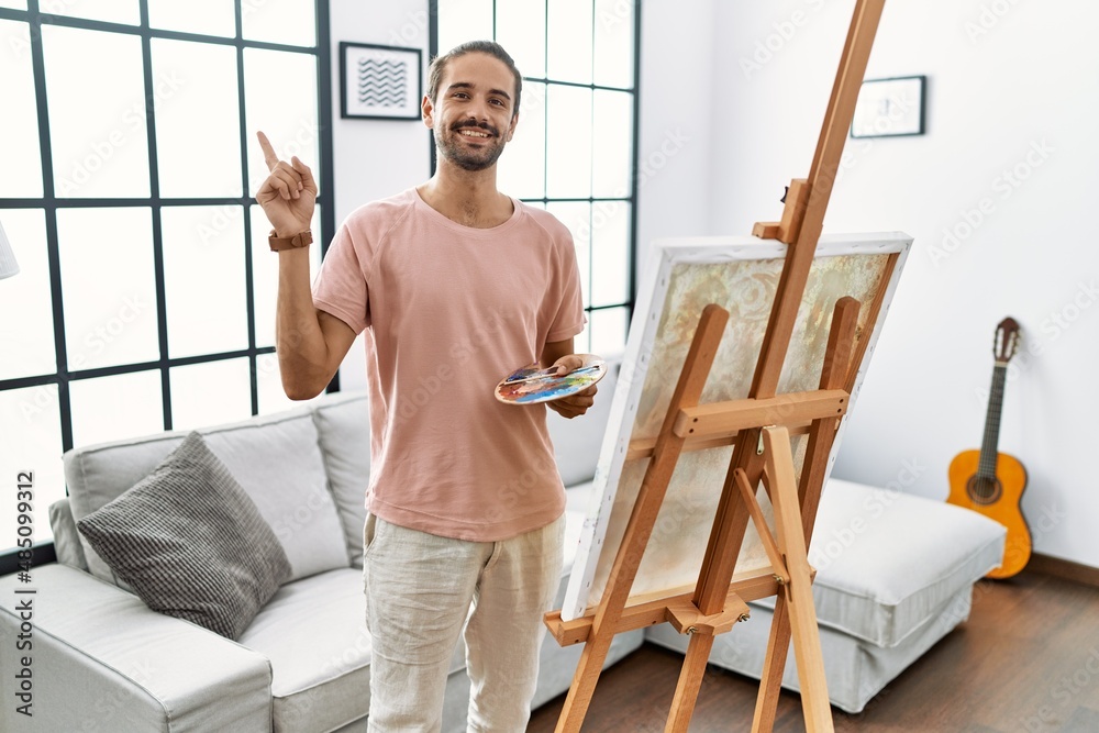 Young hispanic man with beard painting on canvas at home with a big smile on face, pointing with hand finger to the side looking at the camera.