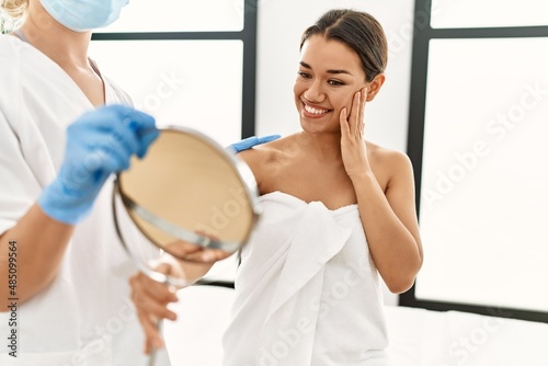 Young latin woman wearing towel looking face on mirror at beauty center
