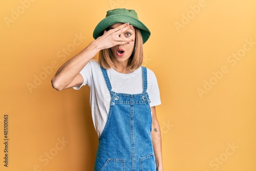 Young caucasian blonde woman wearing denim jumpsuit and hat with 90s style peeking in shock covering face and eyes with hand, looking through fingers with embarrassed expression.